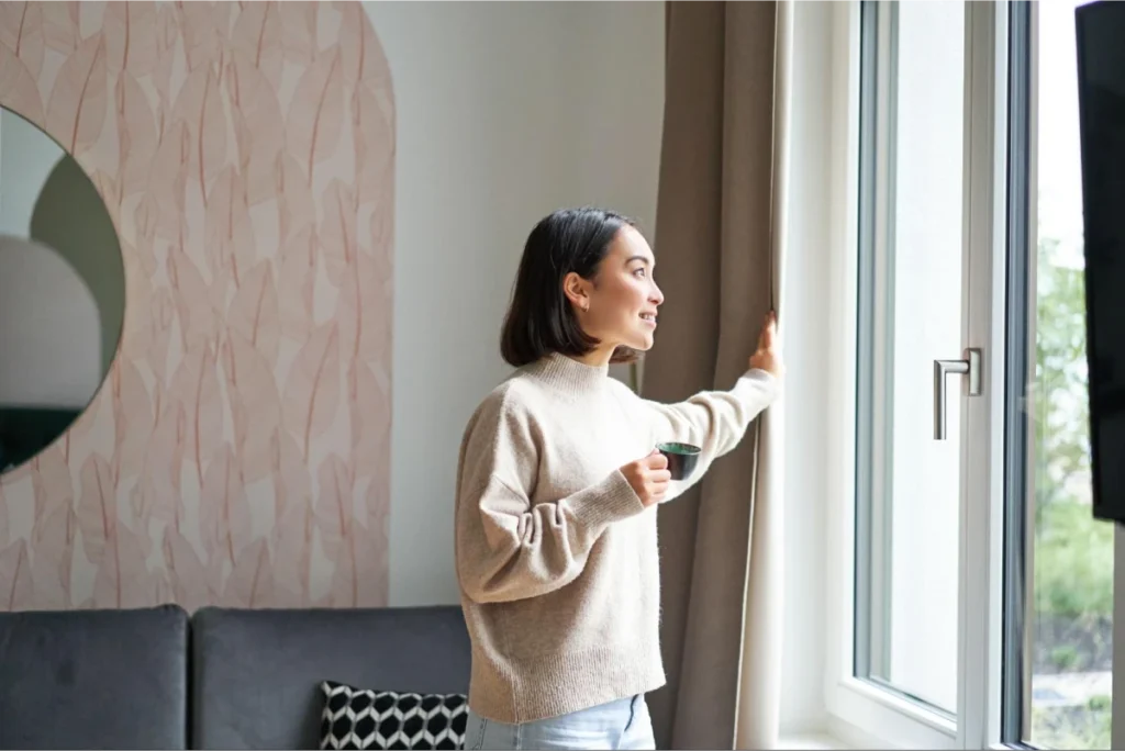 Woman standing by a window holding a coffee cup, smiling and looking outside in a cozy modern living room.