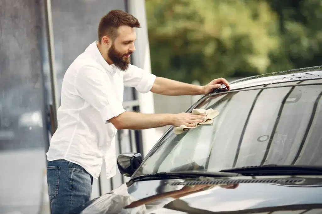Man cleaning a car windshield with a cloth at a car wash on a sunny day.